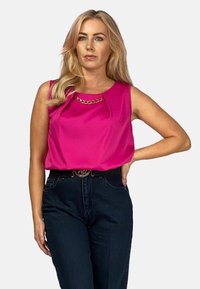 Fuksjowa bluzka na ramiączkach z plisowanym wzorem i złotym łańcuszkiem przy dekolcie, zestawiona z ciemnymi spodniami z wysokim stanem i czarnym paskiem.