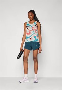 Mulher em roupa desportiva a segurar uma garrafa de água preta, vestindo uma t-shirt sem mangas multicolorida, calças curtas teal, meias brancas e sapatilhas de corrida cor-de-rosa.