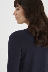 Pull en maille bleu marine avec un col rond, texture douce et design ajusté. Modèle montré de profil, avec des cheveux sombres et ondulés.
