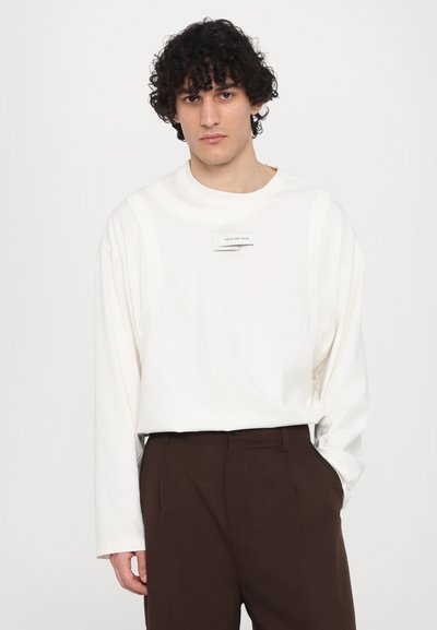 White long-sleeve shirt with a round neckline and textured paneling, paired with dark brown trousers. Features a label on the chest.