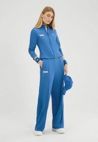 Blauwe sportieve trainingsoutfit met een ritsjack en wijde broekspijpen. Inclusief bijpassende polsbanden en een pet. Zachte textuur met logo-accenten.