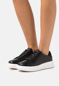 Černé kožené tenisky s hladkým povrchem, kulatou špičkou a bílou gumovou podrážkou. Mají černé tkaničky a minimalistický design.