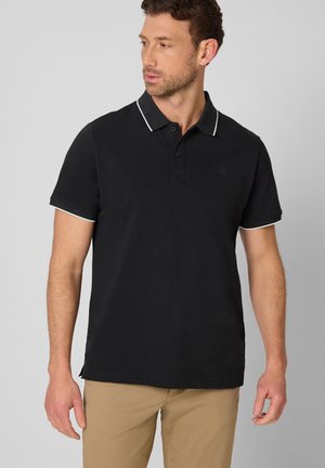 Homme portant un polo noir avec bordure blanche et un pantalon beige, regardant vers sa gauche sur un fond gris uni.