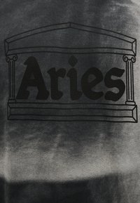 Αρχαιοελληνική δομή ναού με μαύρο περίγραμμα που πλαισιώνει τη λέξη «Κριός» με έντονα, σκούρα γράμματα σε γκρι υφασμένο φόντο.