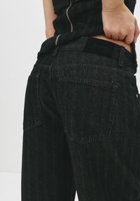 Jeans in denim neri con una texture leggermente sbiadita, dotati di due tasche posteriori e un'etichetta sulla vita. Dettaglio con zip sul lato.