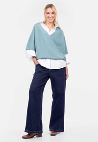 Jasnoniebieski poncho w stylu kopertowym na białej koszuli, połączony z ciemnoniebieskimi szerokimi dżinsami i brązowymi botkami na obcasie, na neutralnym tle.