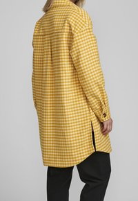Žlutá kostkovaná flanelová košile s rovnými střihy, dlouhými rukávy, bočními rozparky a knoflíkovými manžetami. Měkká textura a viditelný kostkovaný vzor.