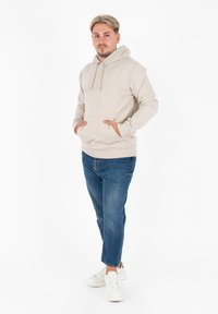 Felpa color beige chiaro con tasca a marsupio e logo ricamato, abbinata a jeans blu e sneakers bianchi, in piedi su uno sfondo neutro.