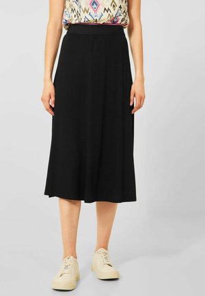 Femme portant une jupe midi noire, un haut sans manches à motif, et des baskets blanches, debout devant un fond gris clair uni.