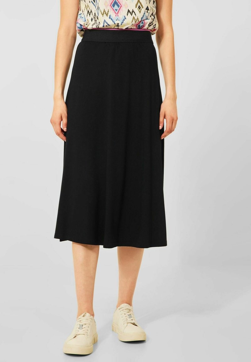 Femme portant une jupe midi noire, un haut sans manches à motif, et des baskets blanches, debout devant un fond gris clair uni.