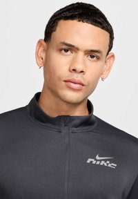 Dunkelgraue, mit Reißverschluss versehene Sportjacke mit Kragen, die eine strukturierte Oberfläche und ein silbernes Nike-Logo auf der linken Brustseite aufweist.