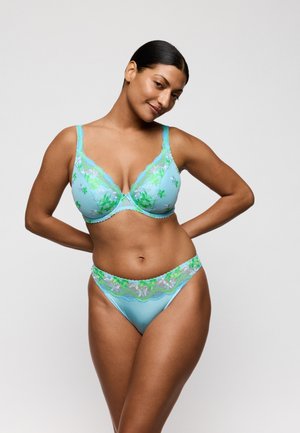 Vrouw die een bijpassend lichtblauw beha- en slipje draagt met groene en witte bloemenkant, poserend met haar handen achter haar rug tegen een effen achtergrond.