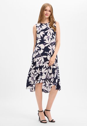 Junge Frau in ärmellosem dunkelblauem Kleid mit großem weißem Blumenmuster, steht mit der Hand am Rock und trägt schwarze Sandalen mit hohen Absätzen.
