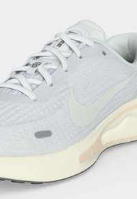 Κοντινή λήψη ενός λευκού παπουτσιού Nike για τρέξιμο με υφή από δίχτυ, λευκά κορδόνια και λεπτομέρεια στη σόλα σε ανοιχτό μπεζ χρώμα.