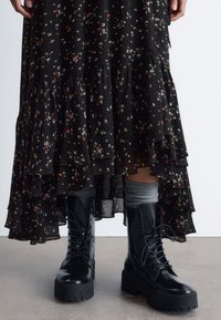 Schwarzes Midikleid mit tiered Rüschen und floralen Mustern, kombiniert mit schwarzen schnürbaren Combat-Stiefeln. Graue Socken sind oberhalb des Knöchels sichtbar. Glatte Stofftextur.