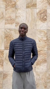 Young man with blond hair smiling, wearing a dark blue quilted jacket and gray pants, standing against a textured beige stone wall.