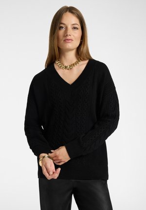 Vrouw draagt een zwarte kabelgebreide V-hals trui, zwarte broek, een chunky gouden ketting en gouden armbanden tegen een effen lichte achtergrond.