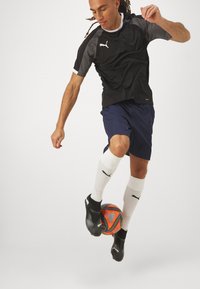 Sort og grå atletisk shirt, marineblå shorts, hvide sokker, sorte fodboldstøvler; spiller jonglerer med en orange fodbold mod en ensfarvet baggrund.