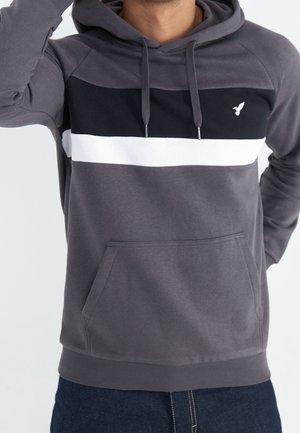 Mężczyzna w szarej bluzie z kapturem z czarno-białymi paskami na piersi i kieszenią typu kangur, ręka uniesiona w pobliżu głowy.