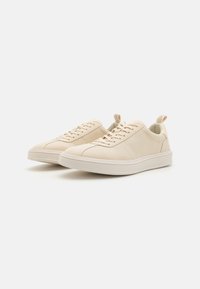 Beige sneakers with a smooth leather upper, round toe, tonal stitching, and a thick rubber sole. Features laces and a rear pull tab.