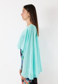 Cappa di chiffon turchese con un design fluido, drappeggio stratificato e una texture trasparente. Indossata sopra un vestito colorato e a fantasia.