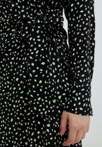 Zwarte jurk met witte abstracte stippen, voorzien van een strik rond de taille en lange mouwen. Gladde stof met een iets losse pasvorm.