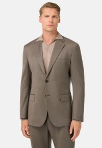 Abito marrone su misura con revers a lancia, chiusura con un solo bottone e due tasche frontali. Tessuto leggero con una sottile texture.