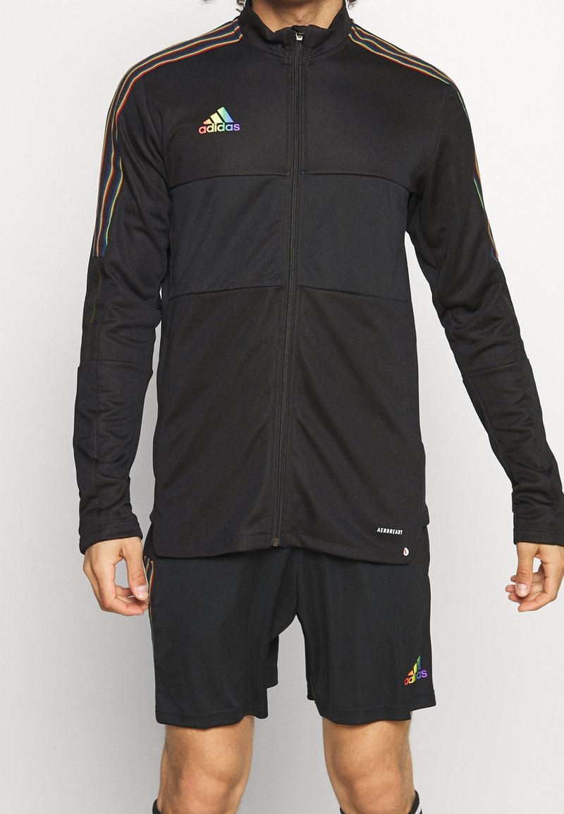 Sort atletisk zip-jakke med regnbue-striber langs ærmerne, med et broderet logo og matchende sorte shorts. Fremstillet af åndbart stof.