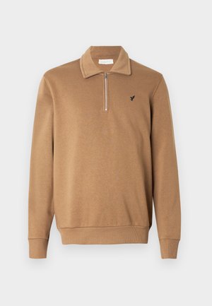 Jersey de pulóver con cuello tipo cremallera en color camel, hecho de tela suave, con dobladillo y puños acanalados, y un pequeño logo negro en el pecho izquierdo.