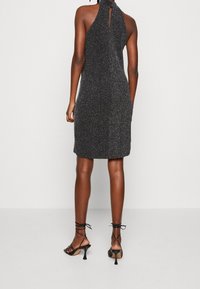 Kvinna klädd i en ärmlös, svart glittrig klänning som når knäna, med detaljer i ryggen i form av en nyckelhålskärning, samt svarta, remmar med hög klack.