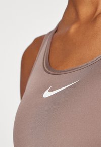 Nike sportski grudnjak u prigušenoj smeđoj tkanini, s izrezom u obliku žlice i istaknutim bijelim swoosh logom sprijeda.