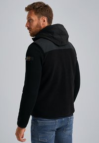 Chaleco con capucha negro hecho de forro polar y nylon. Presenta una textura acanalada, una parte trasera sólida y un parche con el logo en el hombro. Combinado con vaqueros azules.