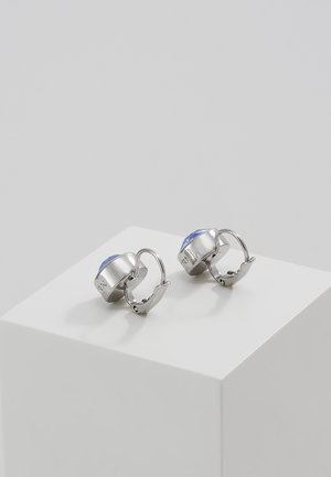 Para srebrnych kolczyków z zapięciem dźwigniowym i okrągłymi jasnoniebieskimi kamieniami, prezentowanych na białej kostce na szarym tle.