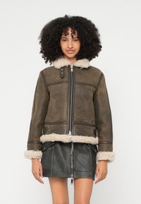 Hnedá bunda zo shearlingu so zipsom vpredu, krémovým golierom a manžetami z umelého kožušina a bočnými vreckami, v kombinácii s čiernou koženou sukňou.