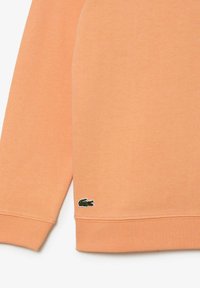 Mouw en onderste gedeelte van een perzikkleurige sweatshirt met een klein groen krokodillogo nabij de zoom op een witte achtergrond.