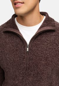 Pullover i mørk mauve, struktureret stof med lodret ribbede. Har en lynlås ved halsen og en kontrastfarvet krave. Båret over en hvid skjorte.