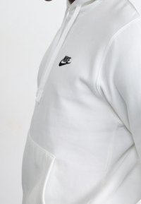 Bílá mikina Nike s kapucí, která má malé černé logo, tkaničku a přední kapsu. Tkanina vypadá hladce a mikina je pohodlně střižená.
