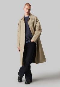 Cappotto lungo beige con colletto e bottoni, indossato sopra un maglione texture blu navy e jeans scuri. Scarpe nere completano il look.
