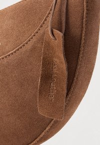 Καφέ προϊόν από σουέντ με καμπύλο σχήμα, με υφή στην επιφάνεια και ανάγλυφη δερμάτινη ετικέτα που προσαρτάται με θηλιά.