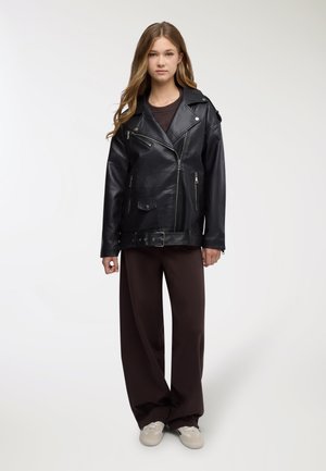 Giacca da motociclista in pelle nera con vita con cintura, tasche con cerniera e ampio rever, abbinata a pantaloni larghi marroni e sneakers.