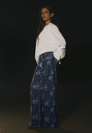 Une femme pose sur un fond sombre, vêtue d’un chemisier blanc ample, d’un long collier à pendentif et d’un jean bleu à motifs et à jambes larges.