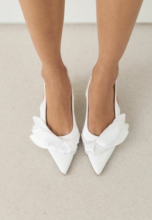 Lábak fehér hegyes orrú lapos cipőkben, nagy díszes szövetmasnikkal, világos színű padlón állva.