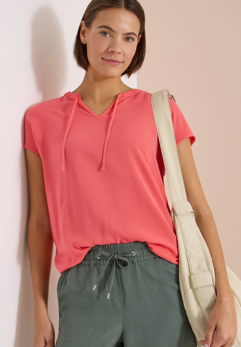 Jeune femme aux cheveux bruns courts porte un haut à capuche corail, un pantalon vert à cordon de serrage et un sac à bandoulière beige, debout contre un mur.