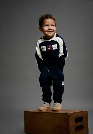 Mørkeblå sweatshirt med hvide ærmer og lapper, parret med matchende joggers. Lysbrune sko med tekstureret design. Står på en trækasse.