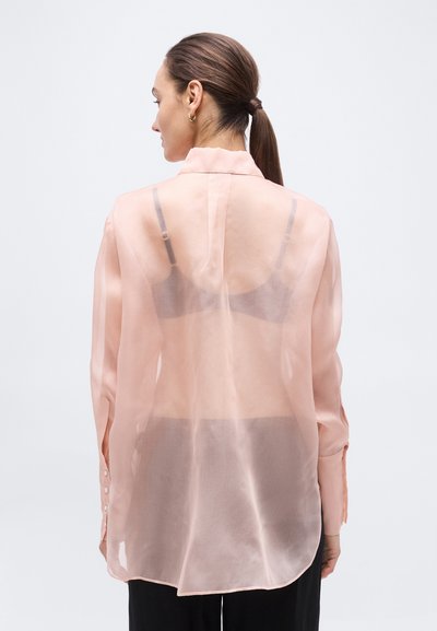 Γυναίκα με σκούρο αλογοουρά, φορά διαφανή ροδακινί μπλούζα, μαύρο σουτιέν και μαύρο παντελόνι, γυρισμένη πλάτη σε απλό φωτεινό φόντο.