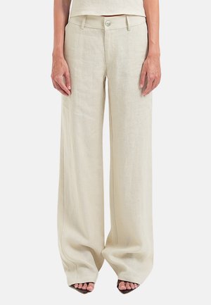 Frau trägt hoch taillierte, weit geschnittene beige Leinenhose mit Knopfverschluss und ärmelloses beiges Oberteil, steht mit entspannten Händen.