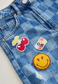 Shorts en denim bleu avec trois patchs brodés : des cerises rouges, un logo coloré "Good Vibes" et un visage souriant jaune. Style texturé et décontracté.