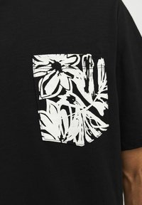 Camiseta negra con un bolsillo texturizado que presenta un estampado floral blanco, mostrando diseños intrincados y colores contrastantes.