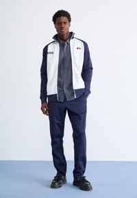 Casual mannenoutfit met een wit en marineblauw kleurgeblokte zipjack, marineblauw geruit shirt, marineblauwe broek en zwarte veterschoenen.