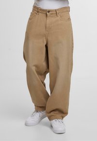 Beige vidde bukser lavet af bomuld, med fem lommer og knappelukning, parret med hvide sneakers på en ensfarvet baggrund.